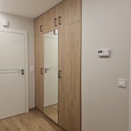 Flormeb - Wysoka szafa wnękowa z lustrem w przedpokoju, wykończona drewnopodobnym laminatem z czarnymi, prostokątnymi uchwytami, widoczna obok białych drzwi z poziomym frezem i drewnianą podłogą.