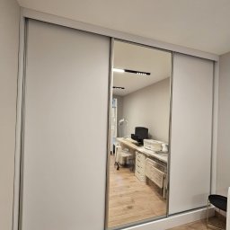 Flormeb - Biała szafa przesuwna z lustrem, w którym odbija się jasne wnętrze gabinetu z biurkiem, krzesłem i oświetleniem punktowym.