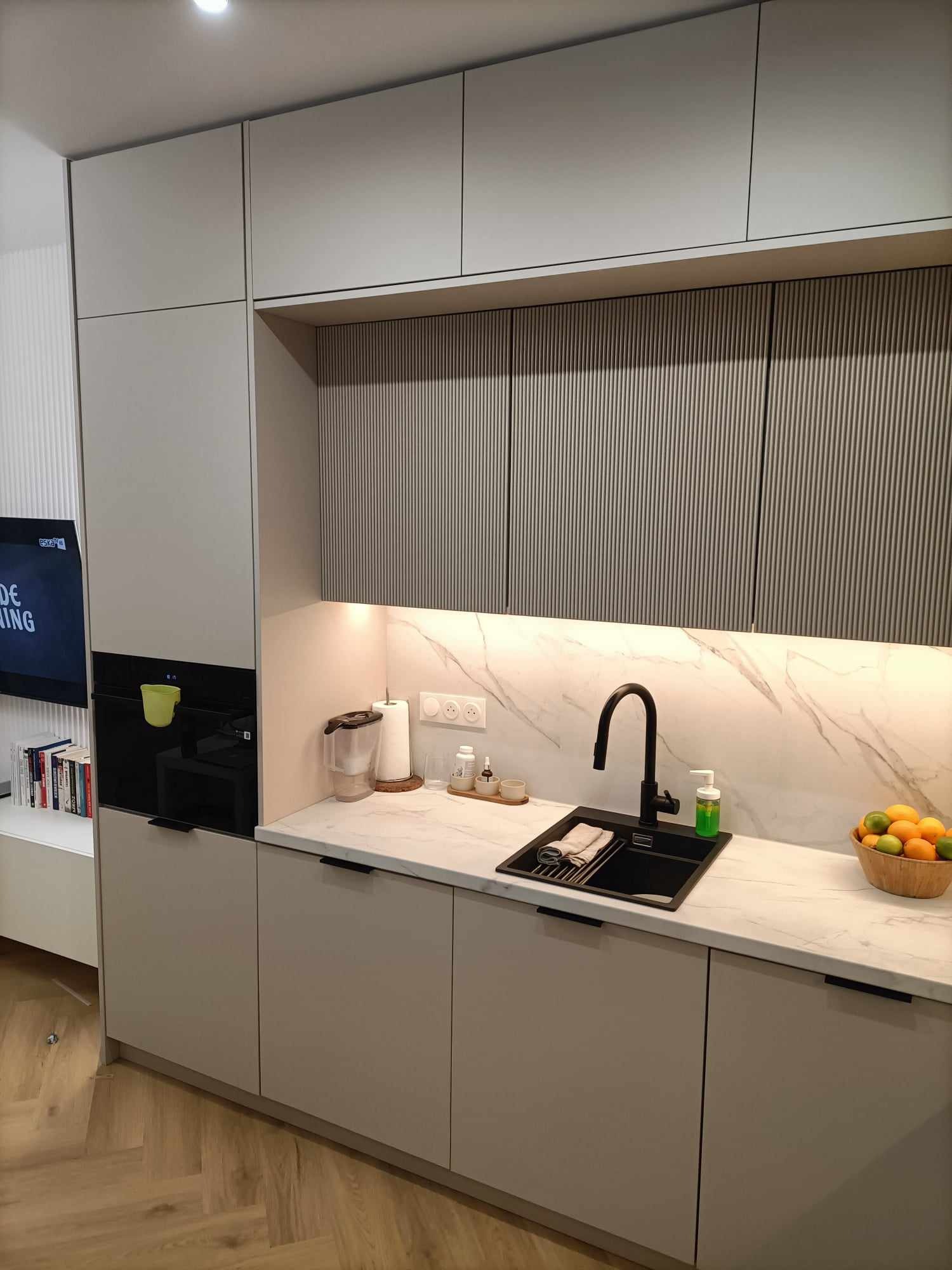 Nowoczesna kuchnia z beżowymi szafkami, marmurowym blatem i czarnym zlewem, oświetlona listwą LED pod górnymi szafkami, z widocznym koszem owoców na blacie.