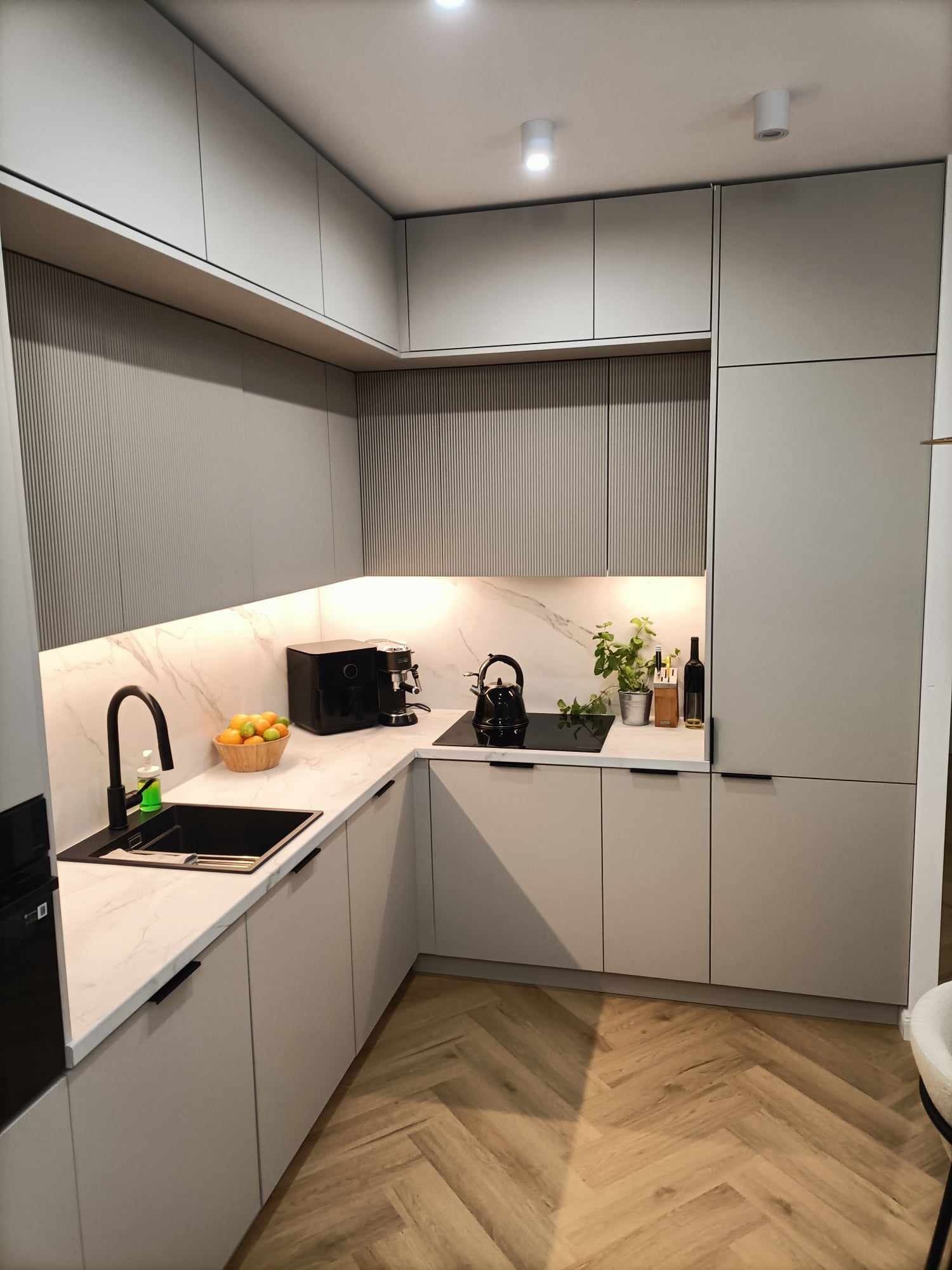 Nowoczesna, jasna kuchnia w Rybnej z szarymi szafkami, marmurowym blatem i czarną armaturą, uzupełniona drewnianą podłogą w jodełkę i dekoracyjnymi elementami.