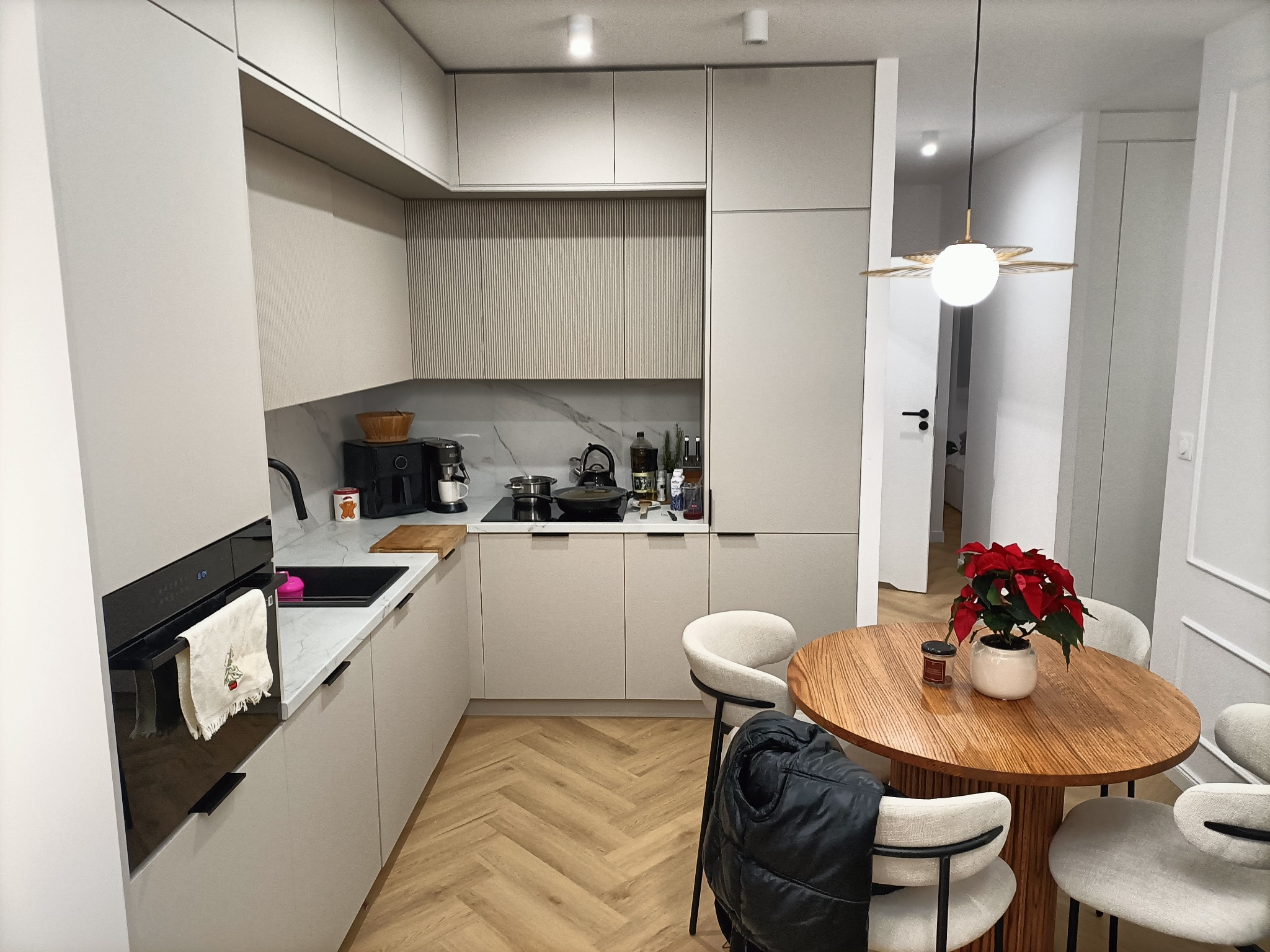 Jasna kuchnia z drewnianym stołem i krzesłami, parkiet w jodełkę, na blacie czajnik, ekspres do kawy i przyprawy, na stole gwiazda betlejemska, na krześle czarna kurtka.