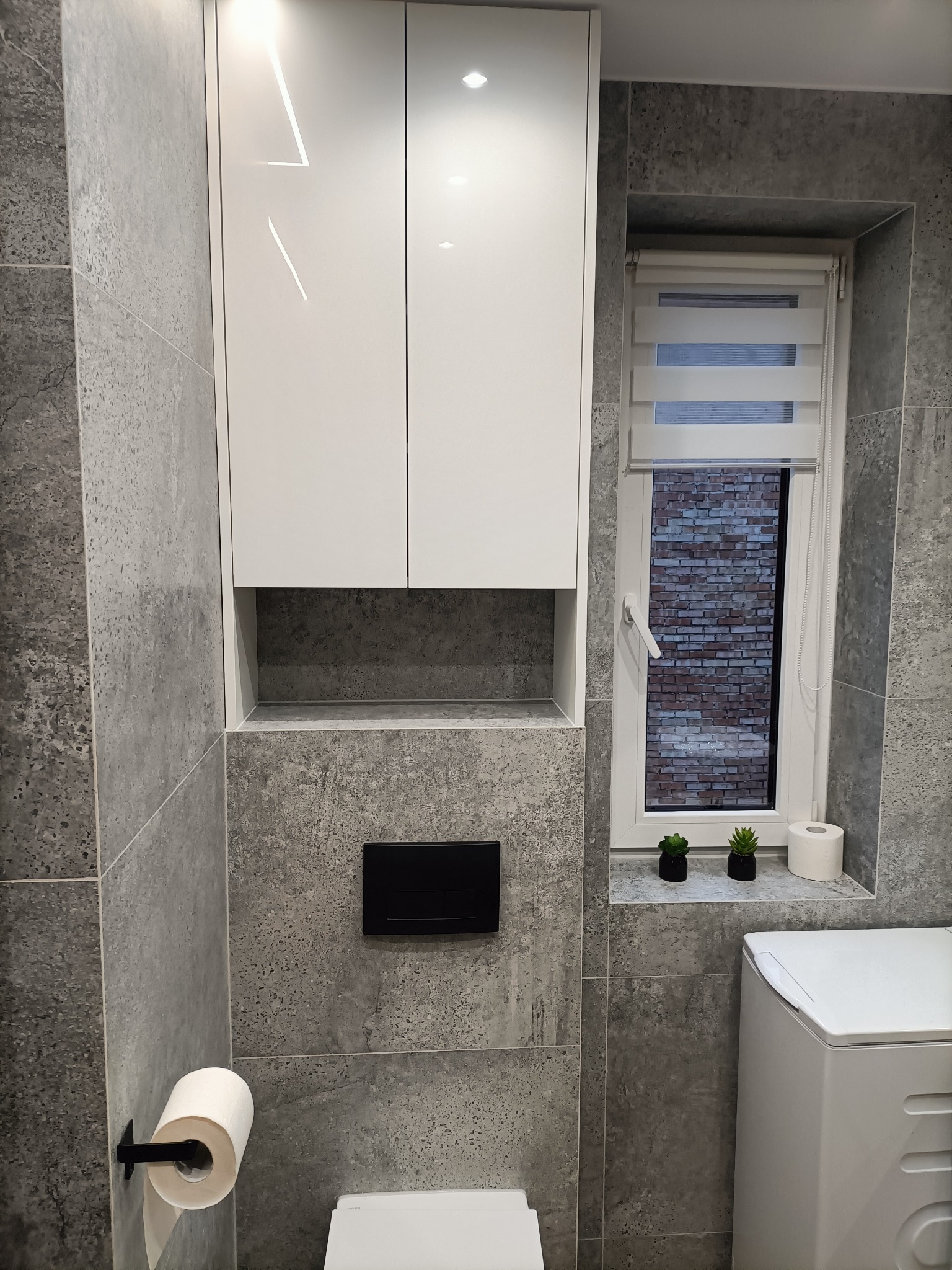 Biała szafka łazienkowa na wymiar z połyskiem, zamontowana nad toaletą w nowoczesnej łazience wykończonej szarą płytką imitującą beton. W tle okno z roletą dzień-noc i dekoracjami.