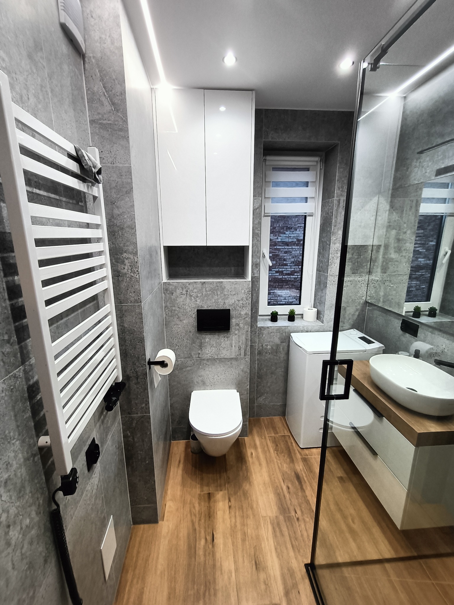 Nowoczesna łazienka z szarymi płytkami, drewnianą podłogą i białymi meblami, w tym szafką nad toaletą, grzejnikiem drabinkowym i umywalką z drewnianym blatem, oświetlona listwami LED.