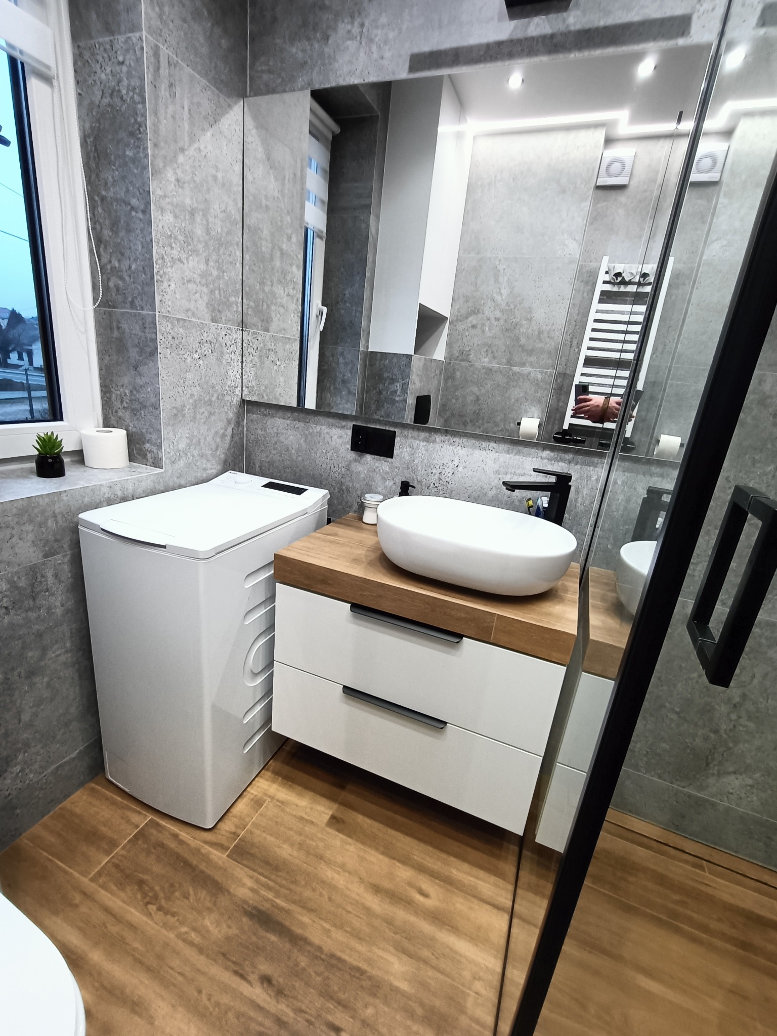 Nowoczesna łazienka z szarą płytką, drewnopodobną podłogą, białą umywalką nablatową na szafce z drewnianym blatem, obok pralka, w tle lustro z oświetleniem i grzejnikiem drabinkowym.