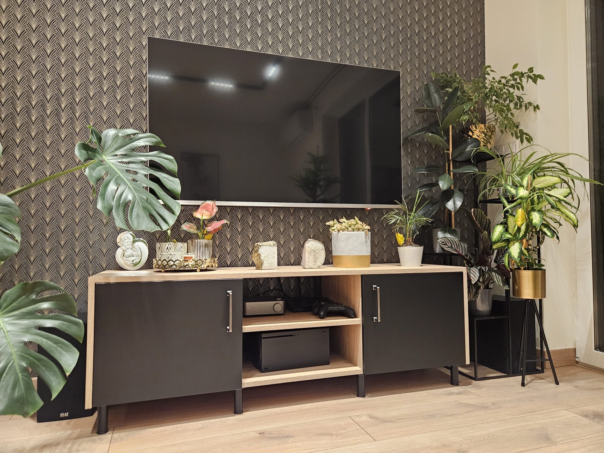 Nowoczesna szafka RTV z czarnymi frontami i drewnianym korpusem, na której stoją rośliny doniczkowe i dekoracje; w tle ściana z tapetą w geometryczny wzór.