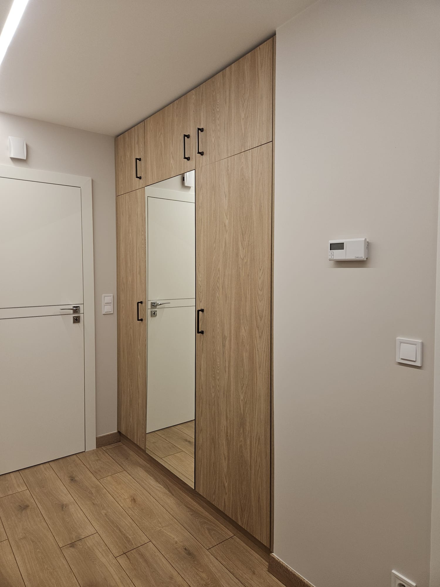 Wysoka szafa wnękowa z lustrem w przedpokoju, wykończona drewnopodobnym laminatem z czarnymi, prostokątnymi uchwytami, widoczna obok białych drzwi z poziomym frezem i drewnianą podłogą.