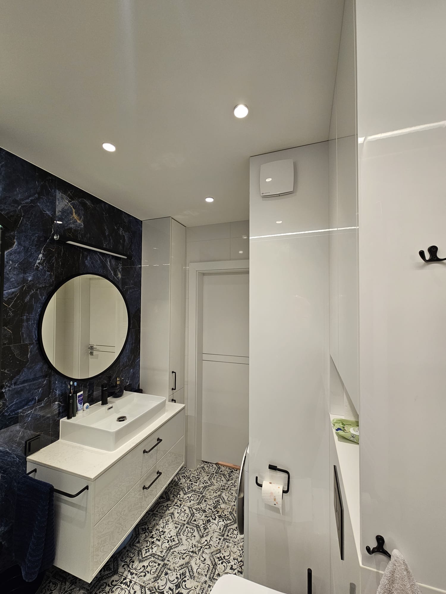 Nowoczesna łazienka z umywalką nablatową na szafce z szufladami, okrągłym lustrem w czarnej ramie na ścianie z efektem ciemnego marmuru, wzorzystą podłogą i białymi ścianami z wbudowanymi szafkami.