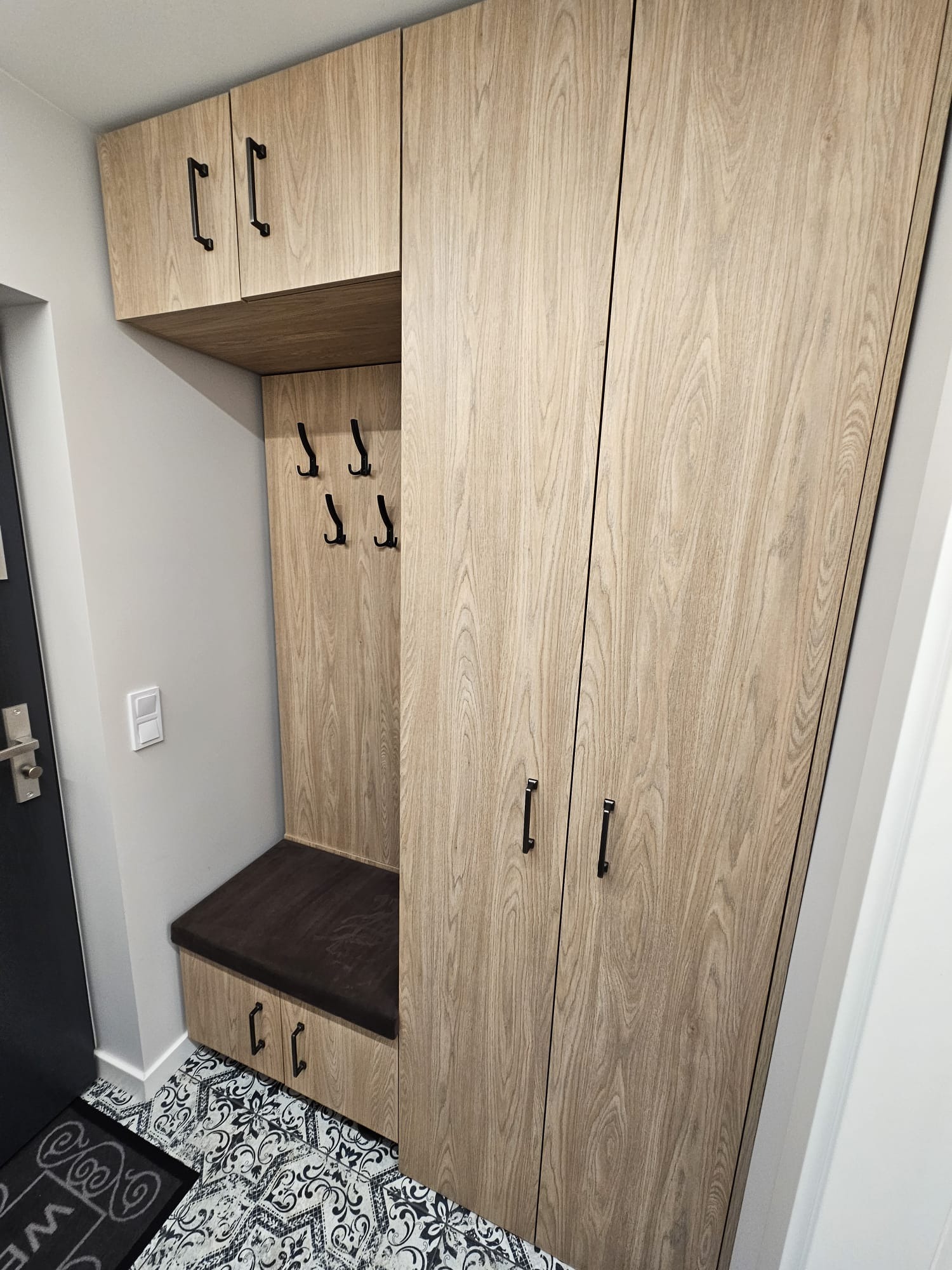 Komplet szaf i siedziska w przedpokoju, wykończone drewnopodobną okleiną z czarnymi uchwytami, na podłodze wzorzyste płytki.