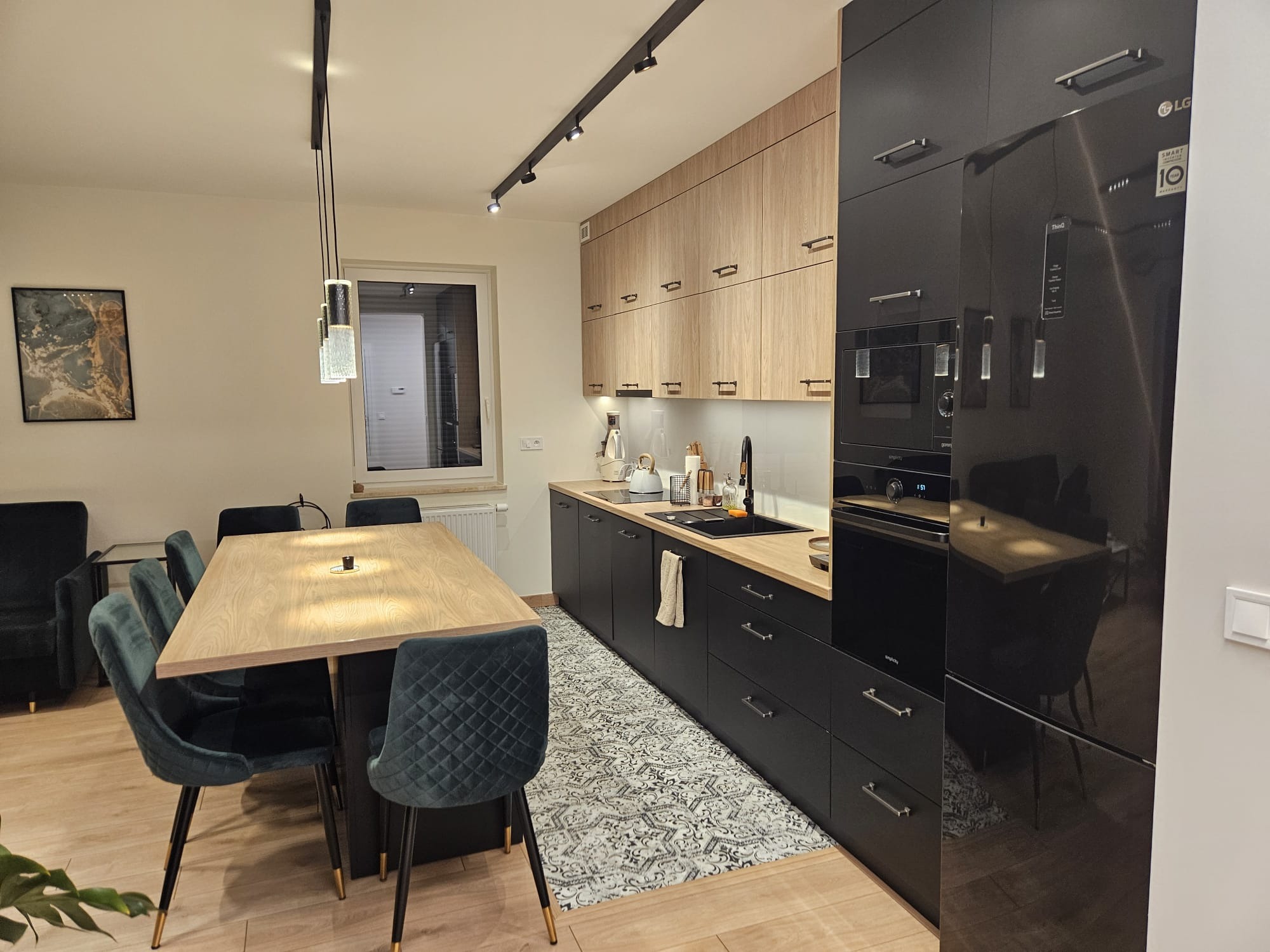 Nowoczesna kuchnia z czarnymi szafkami dolnymi i drewnopodobnymi górnymi, jasnym blatem, czarną lodówką i stołem z zielonymi krzesłami w tle.