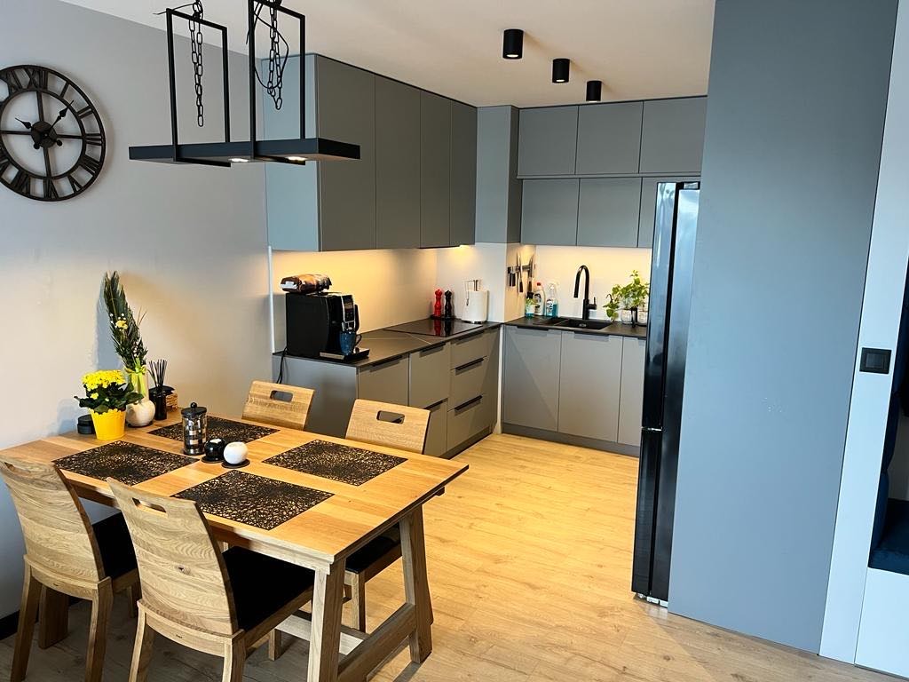 Szara kuchnia na wymiar z drewnianym stołem i krzesłami, czarny zegar na ścianie, nowoczesne oświetlenie sufitowe.