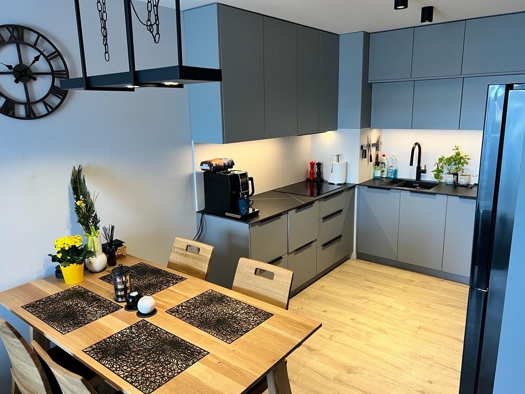 Nowoczesna kuchnia w odcieniach szarości z drewnianym stołem i czarnymi podkładkami, oświetlona punktowymi lampami sufitowymi, z dekoracyjnym zegarem na ścianie.