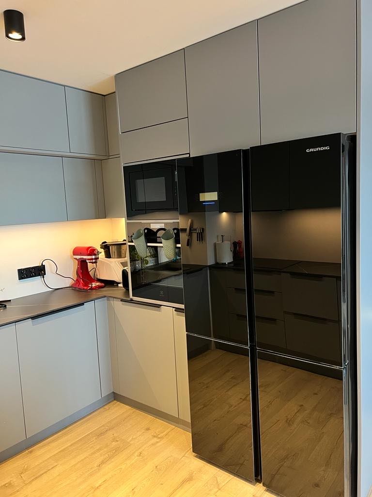 Nowoczesna kuchnia w odcieniach szarości z czarną lodówką side-by-side Grundig, drewnianą podłogą i czerwonym robotem kuchennym na blacie.