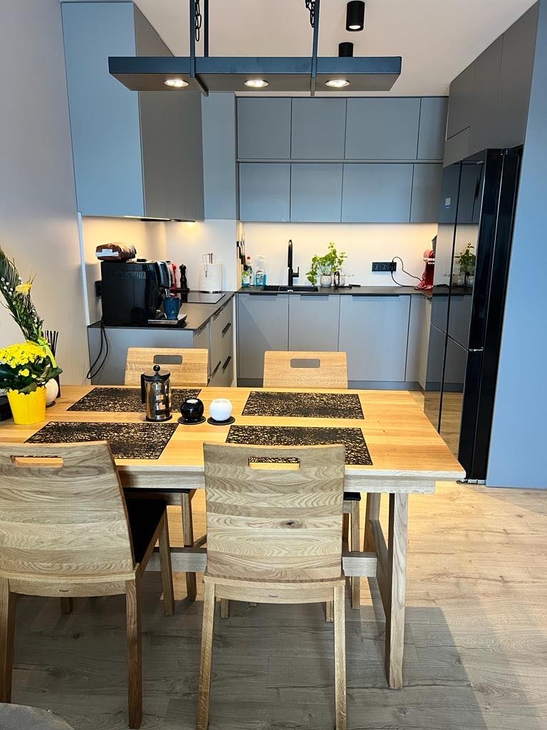 Jasne drewniane krzesła przy stole w nowoczesnej kuchni z szarymi szafkami, czarną lodówką i oświetleniem punktowym.