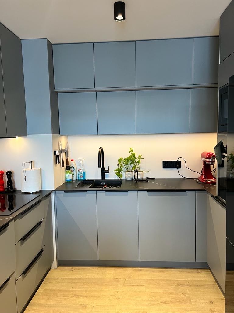 Szara kuchnia z minimalistycznymi frontami, czarnym blatem i armaturą, oświetleniem podszafkowym i drewnianą podłogą. Na blacie widoczny mikser planetarny i doniczka z rośliną.