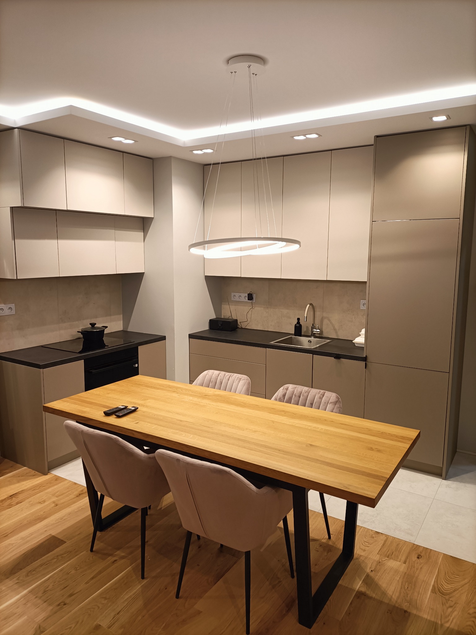 Nowoczesna kuchnia z zabudową w odcieniach beżu, drewnianym stołem z czarnymi nogami i tapicerowanymi krzesłami w kolorze pudrowego różu, oświetlona okrągłą lampą wiszącą.