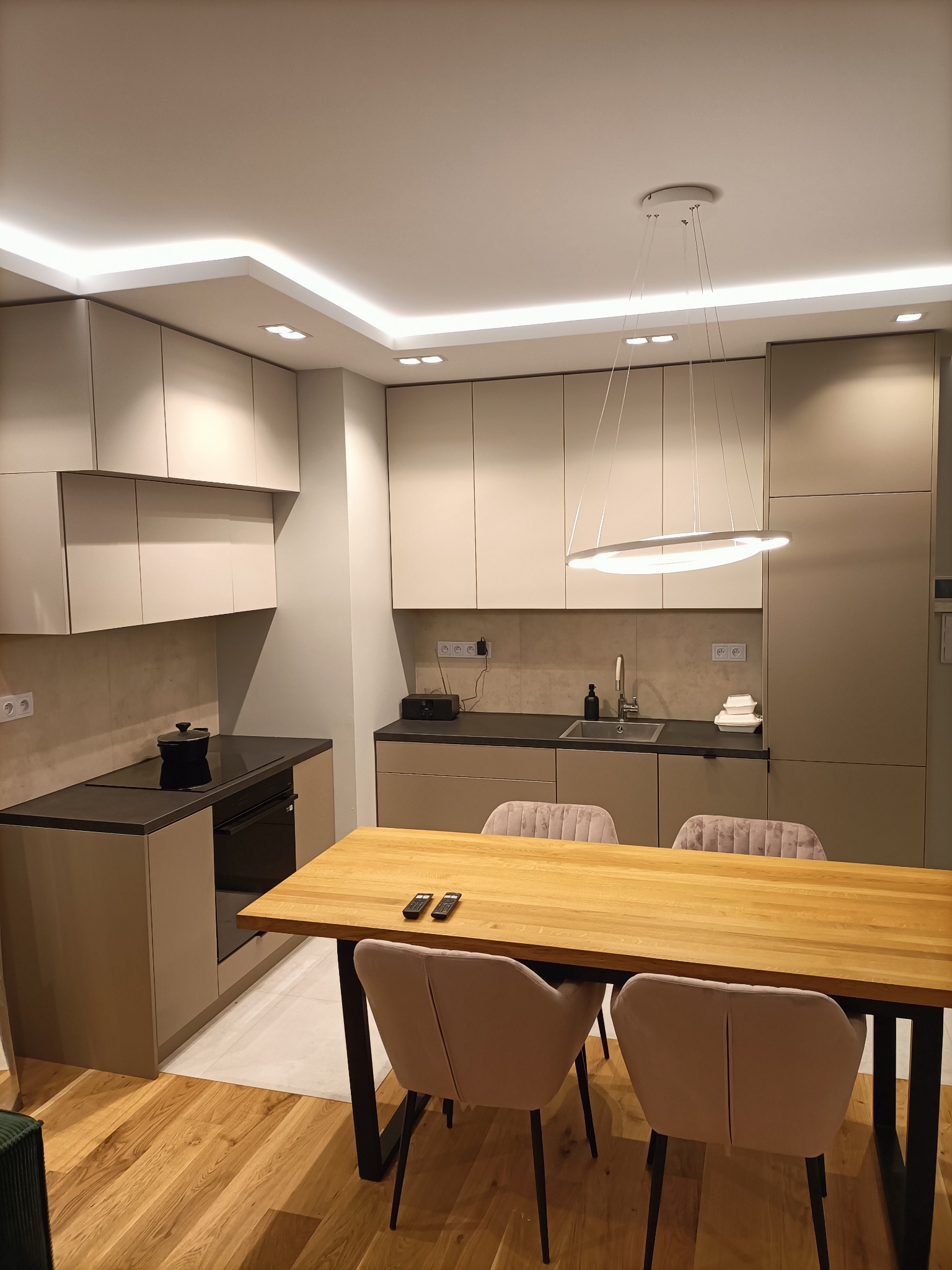 Nowoczesna kuchnia z beżowymi szafkami, czarnym blatem i drewnianym stołem z krzesłami w kolorze pudrowego różu, oświetlona ledowym światłem pod sufitem.