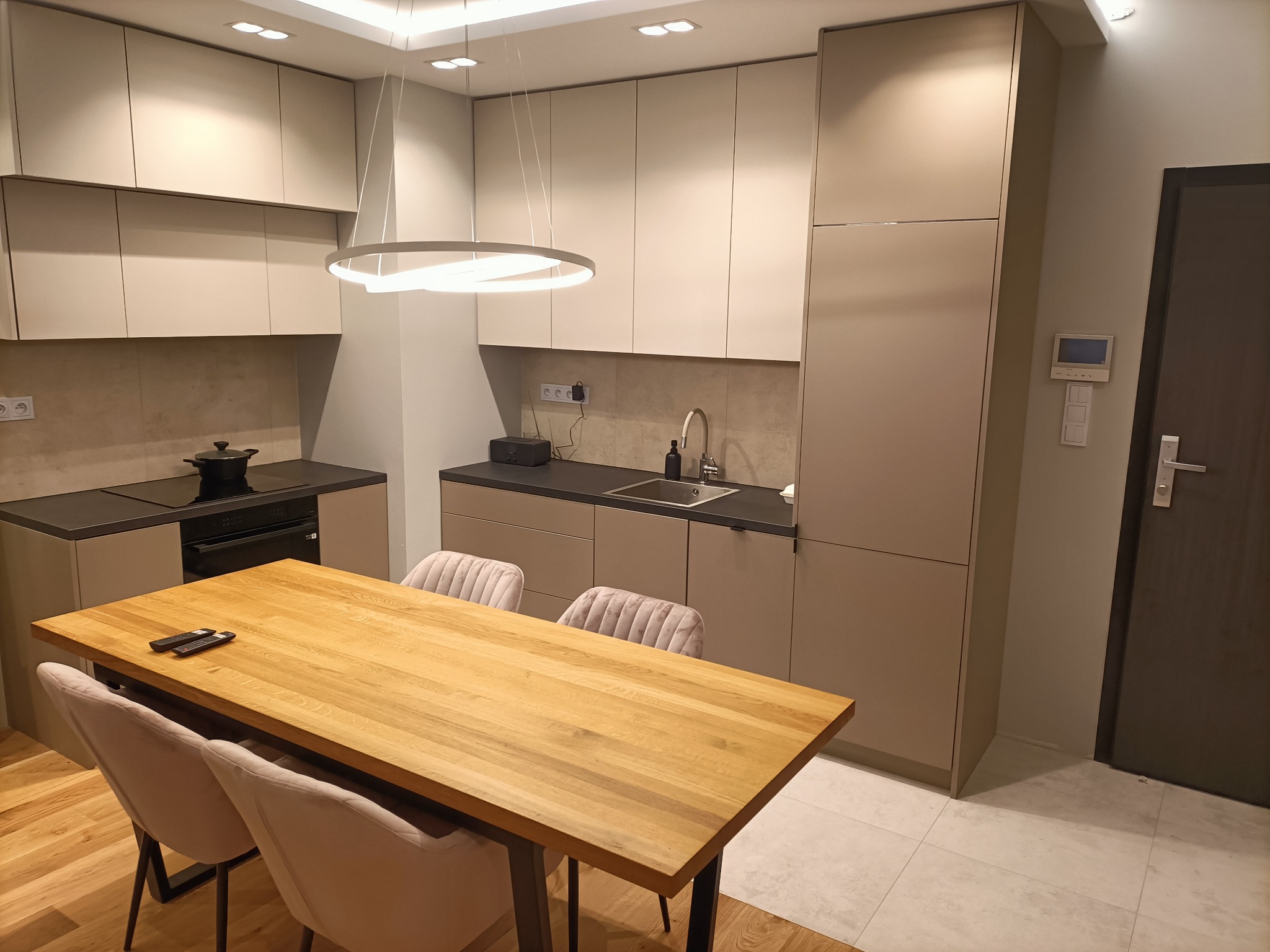 Nowoczesna kuchnia z beżowymi szafkami, czarnym blatem, drewnianym stołem z czterema krzesłami i okrągłą lampą wiszącą