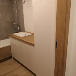 Nowoczesna łazienka z białą szafką pod umywalką z drewnianym blatem, widoczna bateria prysznicowa i fragment drewnianej podłogi.