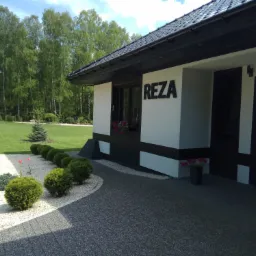 Elewacja nowoczesnego domu z czarnym napisem REZA, zadbany trawnik i ozdobne krzewy