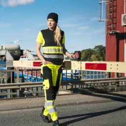 Kobieta w czarno-żółtym komplecie ubrań z elementami odblaskowymi, stojąca na tle mostu zwodzonego i czerwonego budynku z metalową drabinką.