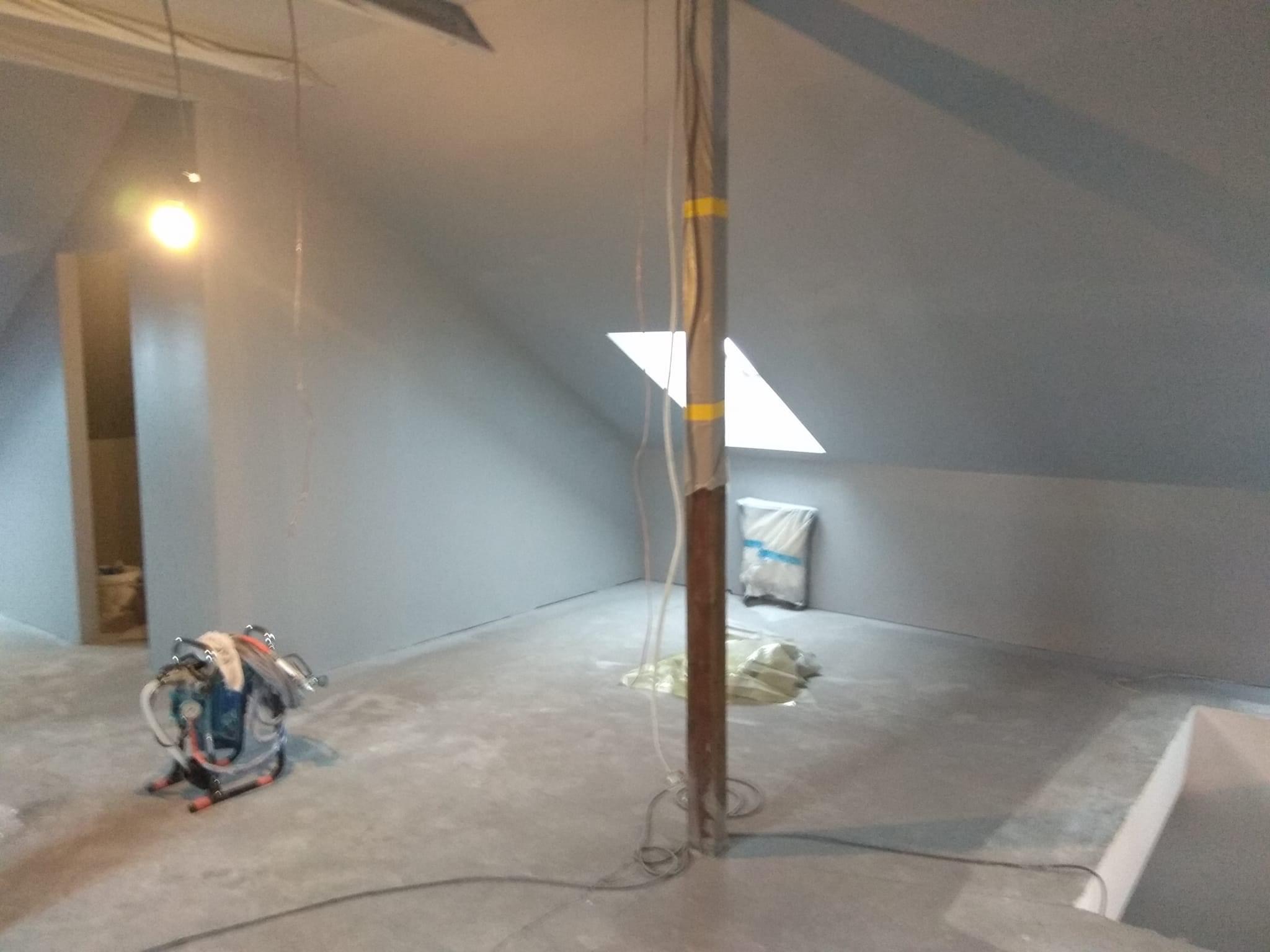 Poddasze po szpachlowaniu i malowaniu na szaro, widoczny sprzęt do malowania, zabezpieczona folią paczka, okno dachowe i pionowa rura z taśmą ostrzegawczą.