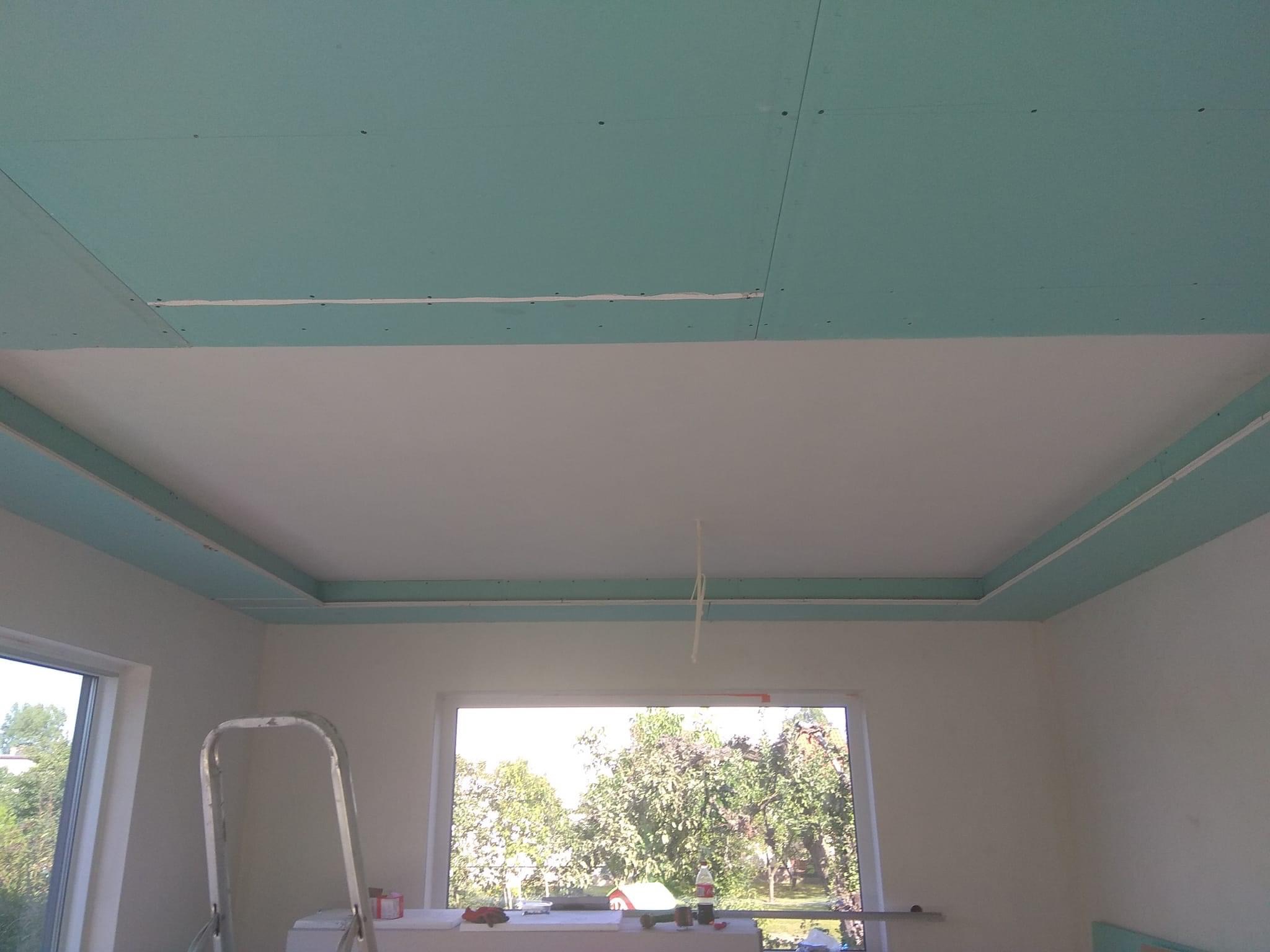 Niedokończony sufit podwieszany z płyty gipsowo-kartonowej, widoczne łączenia i śruby, w tle okno z widokiem na drzewa, drabina oparta o ścianę.