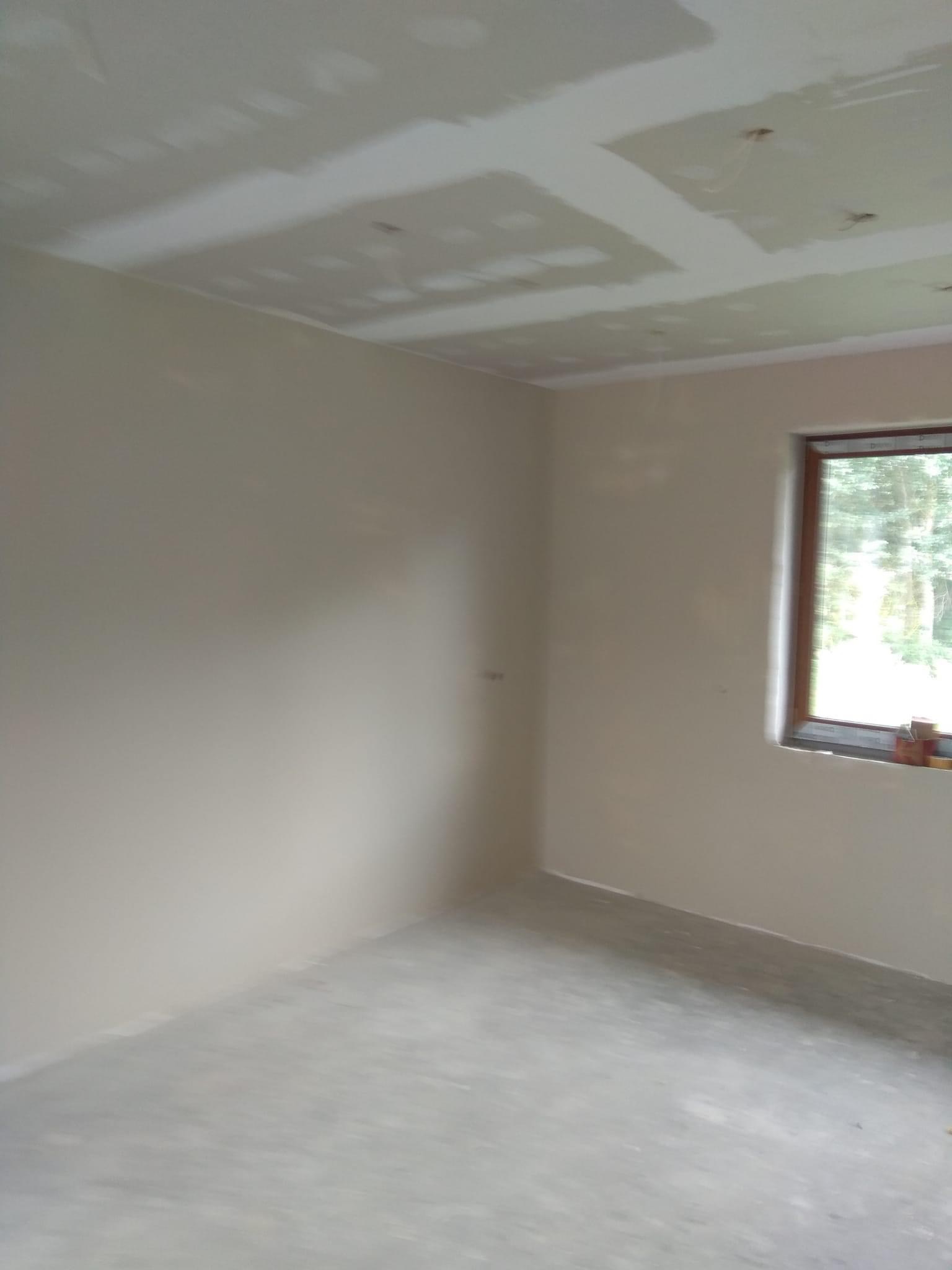 Pokój w trakcie prac wykończeniowych, ściany i sufit po szpachlowaniu, widoczne niedoskonałości i nierówności, surowy beton na podłodze, drewniane okno z widokiem na zieleń.