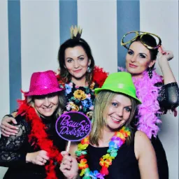 Cztery uśmiechnięte kobiety pozują do zdjęcia w fotobudce, ubrane w kolorowe kapelusze, boa z piór i zabawne okulary, trzymając tabliczkę z napisem 'Baw się dobrze' na tle w biało-szare pasy.