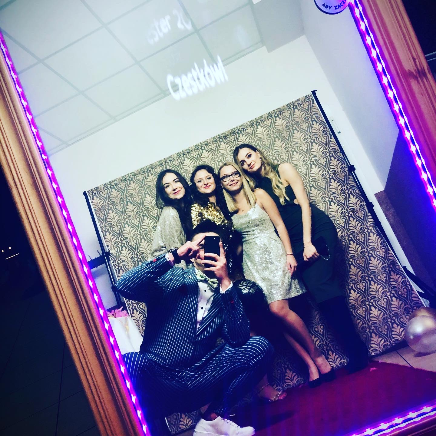 Grupa czterech kobiet pozuje do zdjęcia w fotobudce z lustrzaną ramą podświetloną fioletowymi diodami, mężczyzna w pinstripe garniturze robi im zdjęcie telefonem, w tle wzorzysta ścianka i napis...