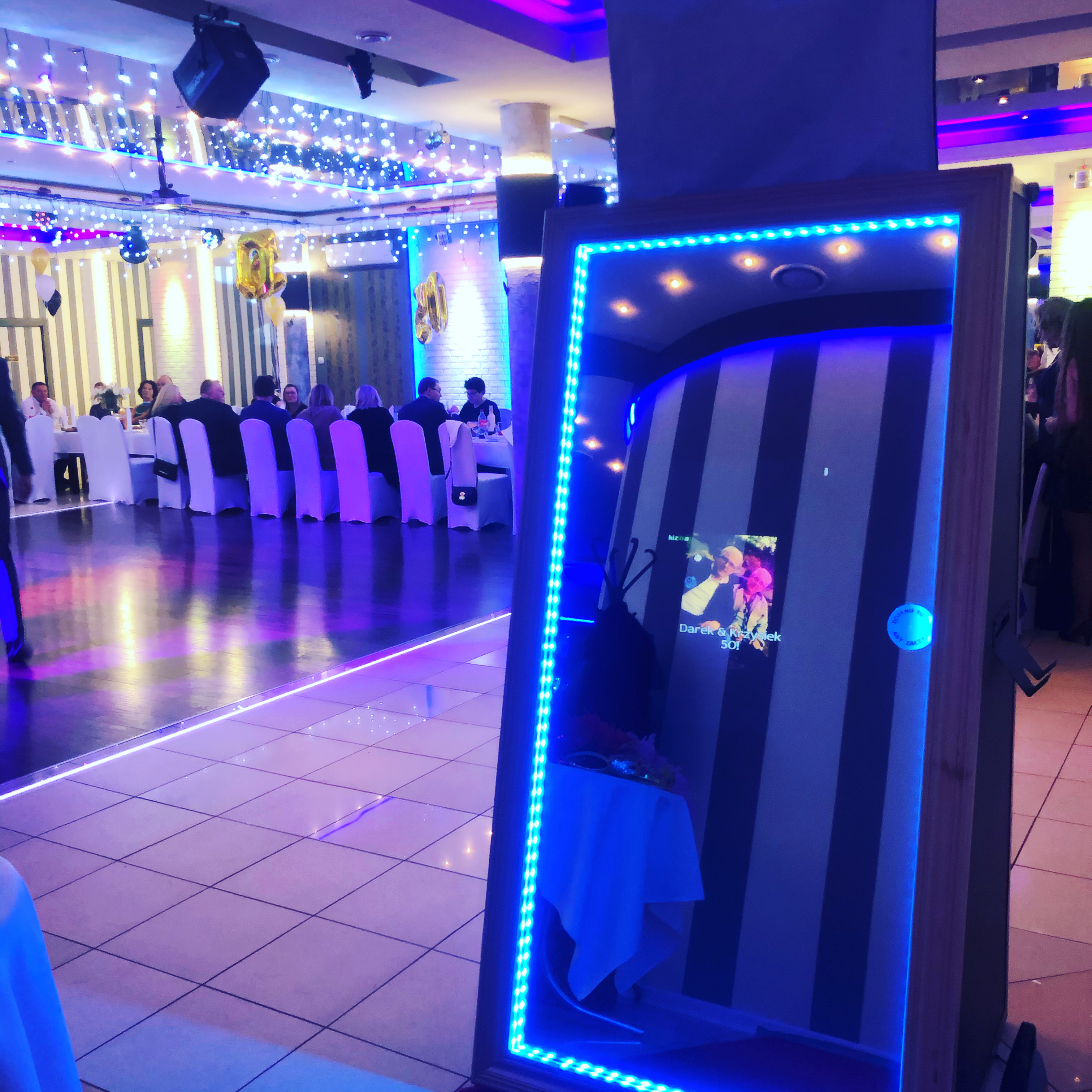 Fotobudka w formie lustra z niebieskim oświetleniem LED na sali balowej podczas imprezy, w tle widoczni goście przy stole.