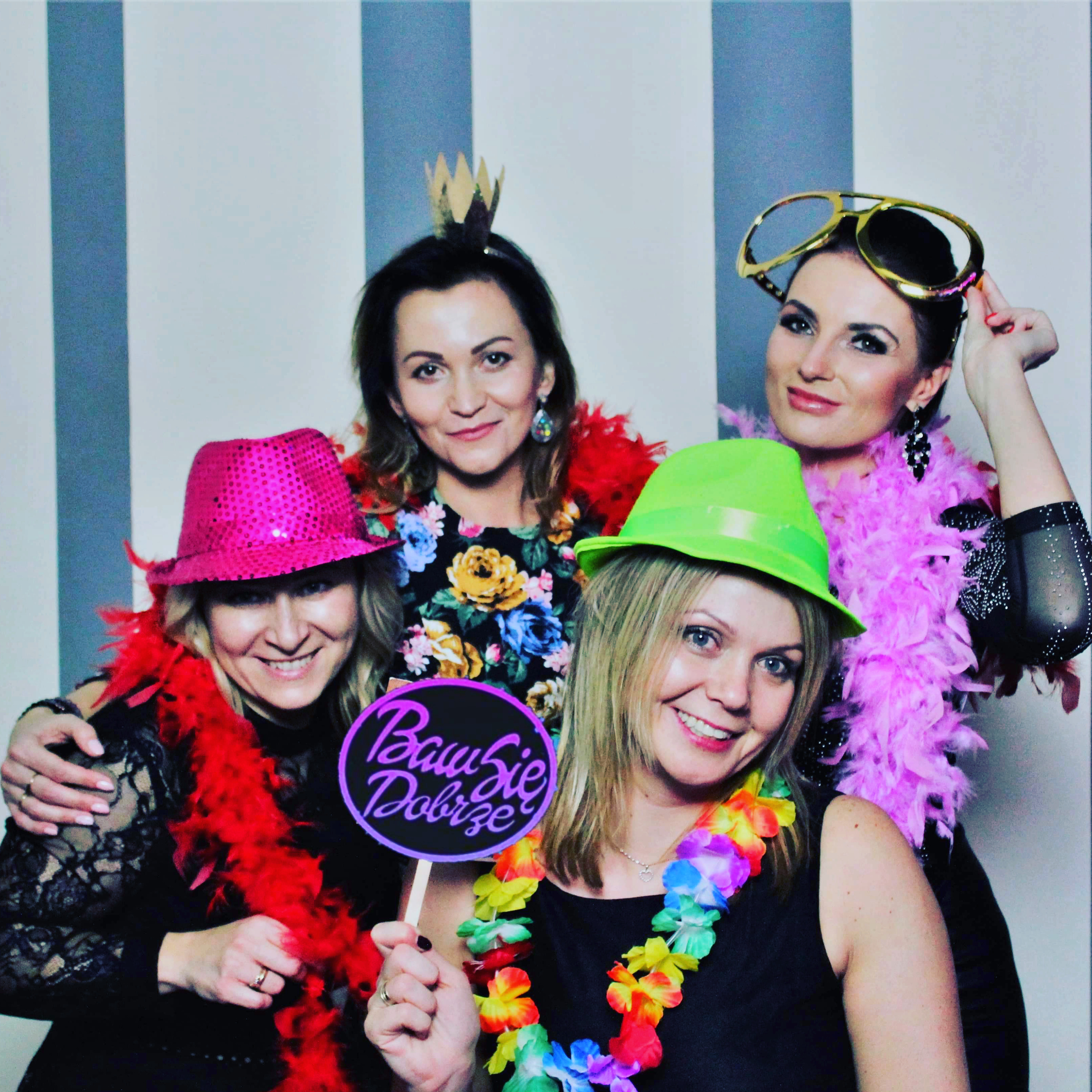 Cztery uśmiechnięte kobiety pozują do zdjęcia w fotobudce, ubrane w kolorowe kapelusze, boa z piór i zabawne okulary, trzymając tabliczkę z napisem 'Baw się dobrze' na tle w biało-szare pasy.