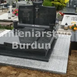 Czarny, podwójny pomnik z krzyżem i napisem 'Ave Maria', otoczony szarą kostką brukową na cmentarzu.