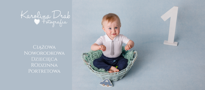 Radosny roczny chłopiec w koszyku z zielonym futerkiem, ubrany w białe body z szarym krawatem i granatowe spodnie, obok drewniana cyfra jeden i dekoracja w kształcie autka na błękitnym tle.