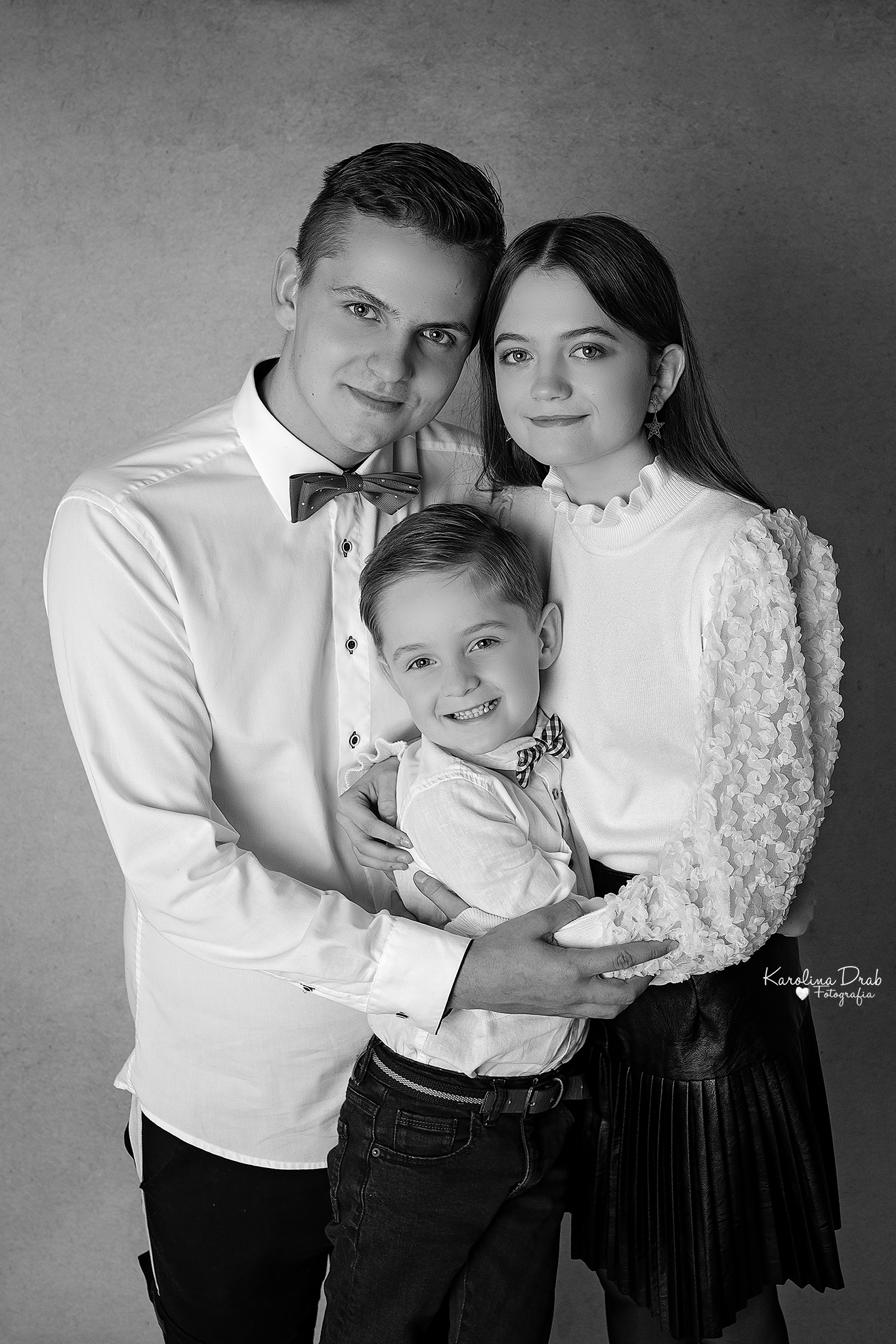 Czarno-białe portretowe ujęcie rodziny: ojca, matki i syna, wszyscy ubrani w eleganckie stroje, uśmiechnięci, na jednolitym tle.