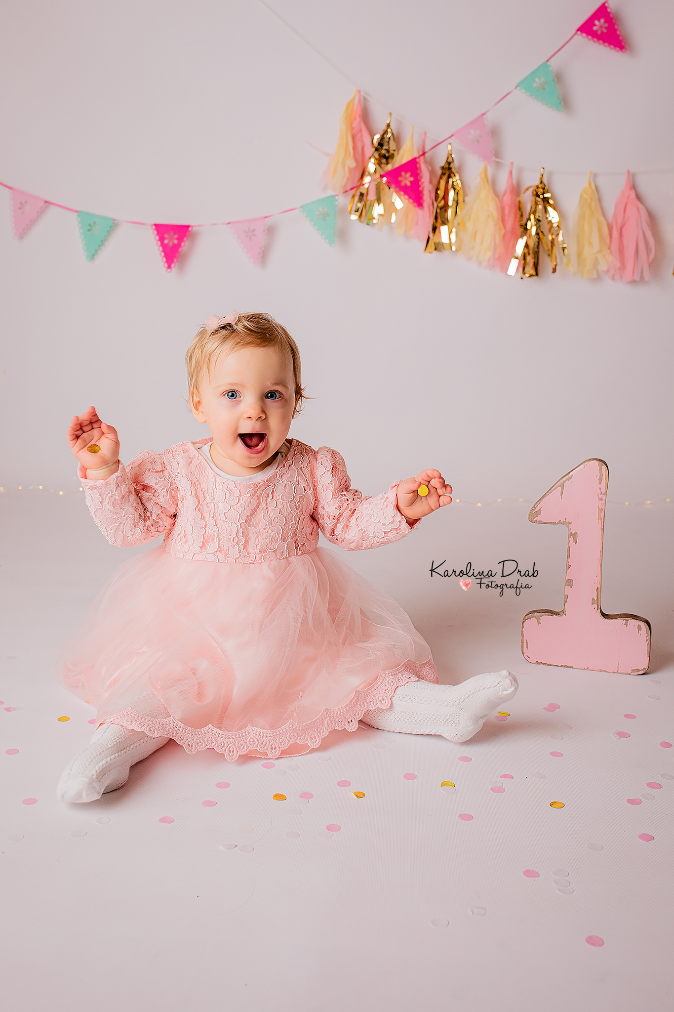 Radosna roczna dziewczynka w różowej sukience siedzi na białym tle z konfetti, trzymając w dłoniach złote kółeczka, obok stoi duża, różowa cyfra '1', a w tle wisi kolorowa girlanda.