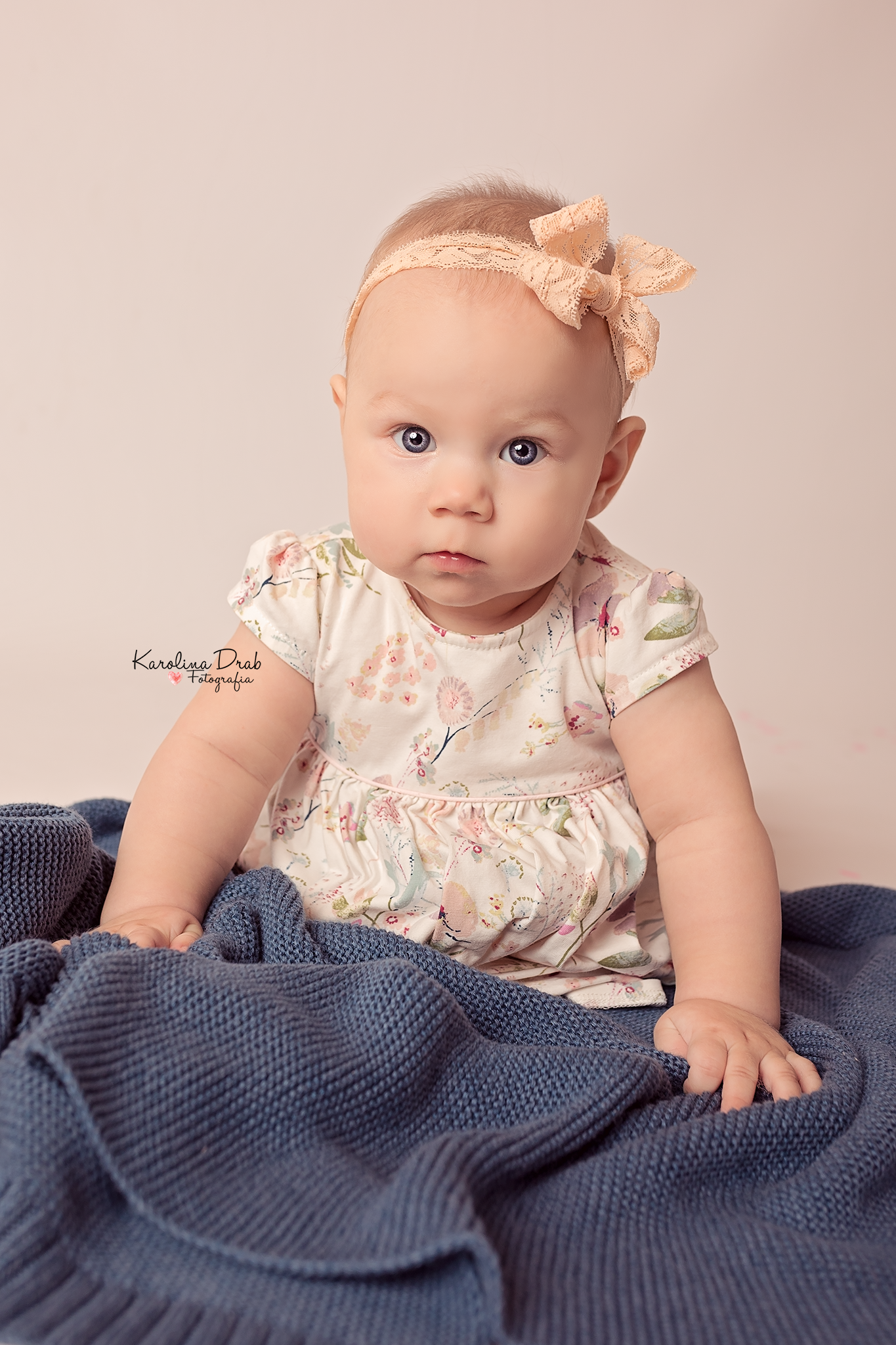 Niemowlę z niebieskimi oczami, ubrane w kwiecistą sukienkę i koronkową opaskę z kokardą, siedzi na niebieskim, dzierganym kocu na jasnym tle.