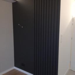 Tomek - Nowoczesna ściana w pokoju z pionowymi, czarnymi panelami dekoracyjnymi przylegającymi do gładkiej, czarnej ściany i białej ściany, widoczne gniazdka elektryczne i wystające kable.