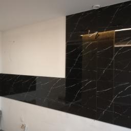 Tomek - Częściowo wykończona ściana z czarnych, błyszczących płytek imitujących marmur z białymi żyłami, kontrastująca z białą, niepomalowaną ścianą w nowym mieszkaniu.