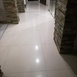 Tomek - Błyszcząca, beżowa podłoga z płytek w korytarzu, z kolumnami obłożonymi kamieniem dekoracyjnym po bokach, odbicie w lustrzanej szafie w tle.