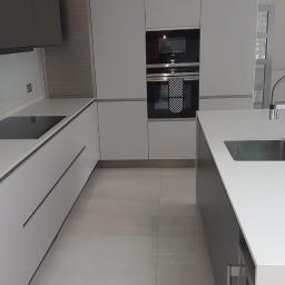 Tomek - Nowoczesna kuchnia w Legionowie z białymi i szarymi szafkami, czarną płytą grzewczą i piekarnikiem, minimalistycznym oświetleniem oraz jasną podłogą z płytek.