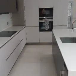 Nowoczesna kuchnia w Legionowie z białymi i szarymi szafkami, czarną płytą grzewczą i piekarnikiem, minimalistycznym oświetleniem oraz jasną podłogą z płytek.