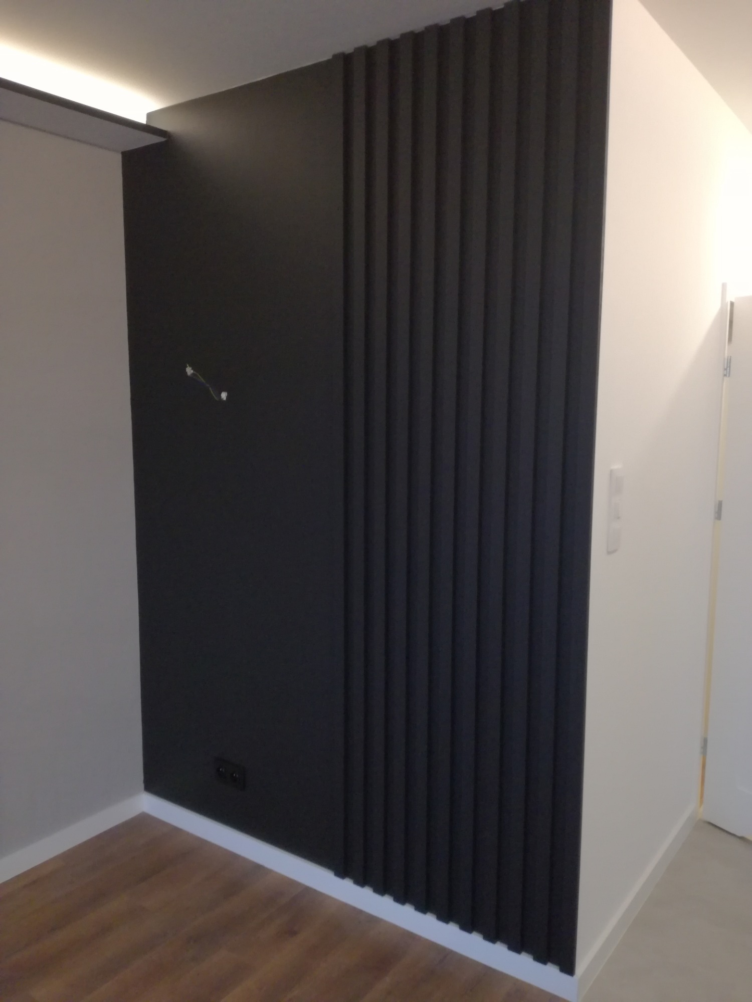 Nowoczesna ściana w pokoju z pionowymi, czarnymi panelami dekoracyjnymi przylegającymi do gładkiej, czarnej ściany i białej ściany, widoczne gniazdka elektryczne i wystające kable.
