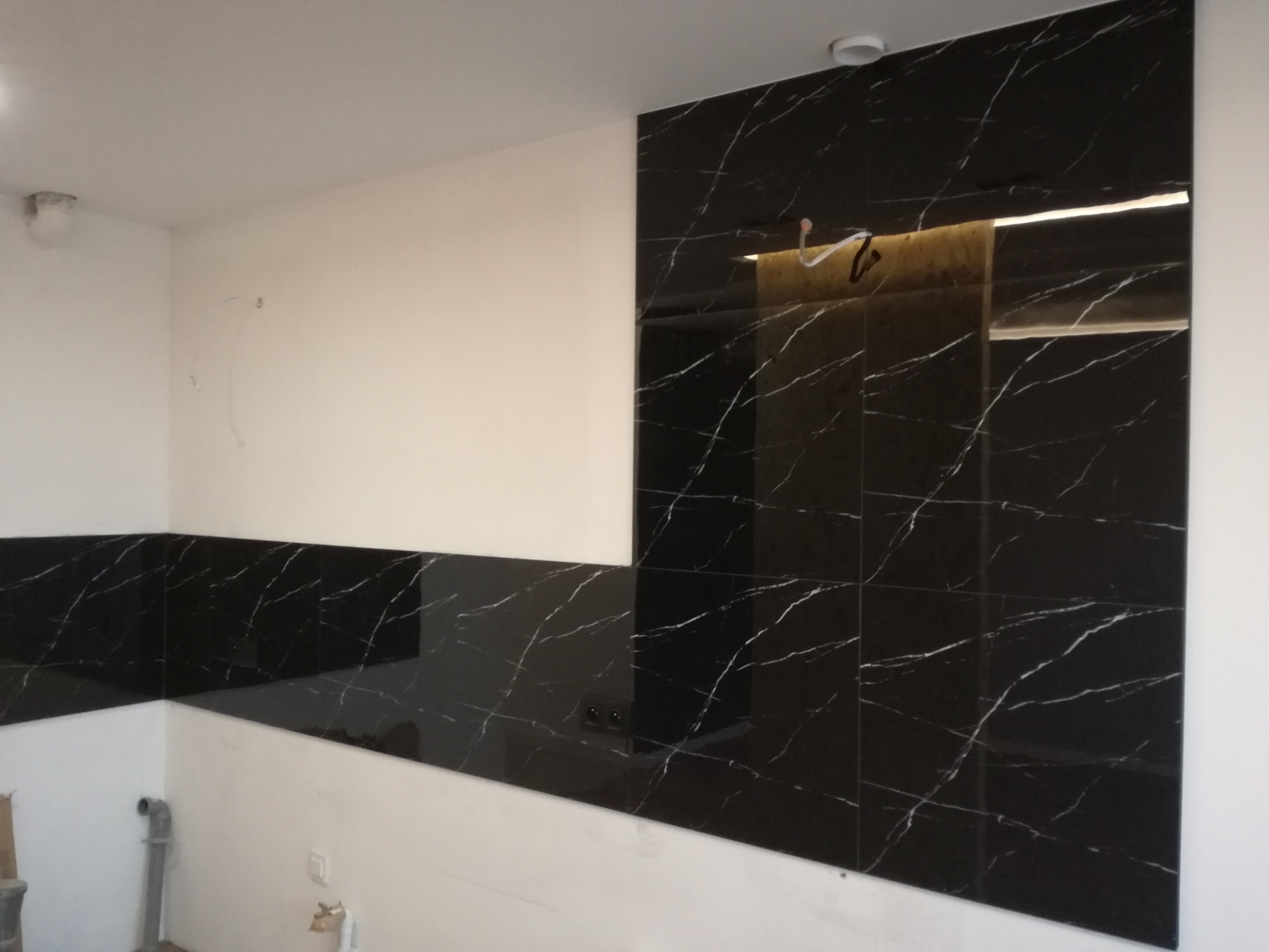 Częściowo wykończona ściana z czarnych, błyszczących płytek imitujących marmur z białymi żyłami, kontrastująca z białą, niepomalowaną ścianą w nowym mieszkaniu.