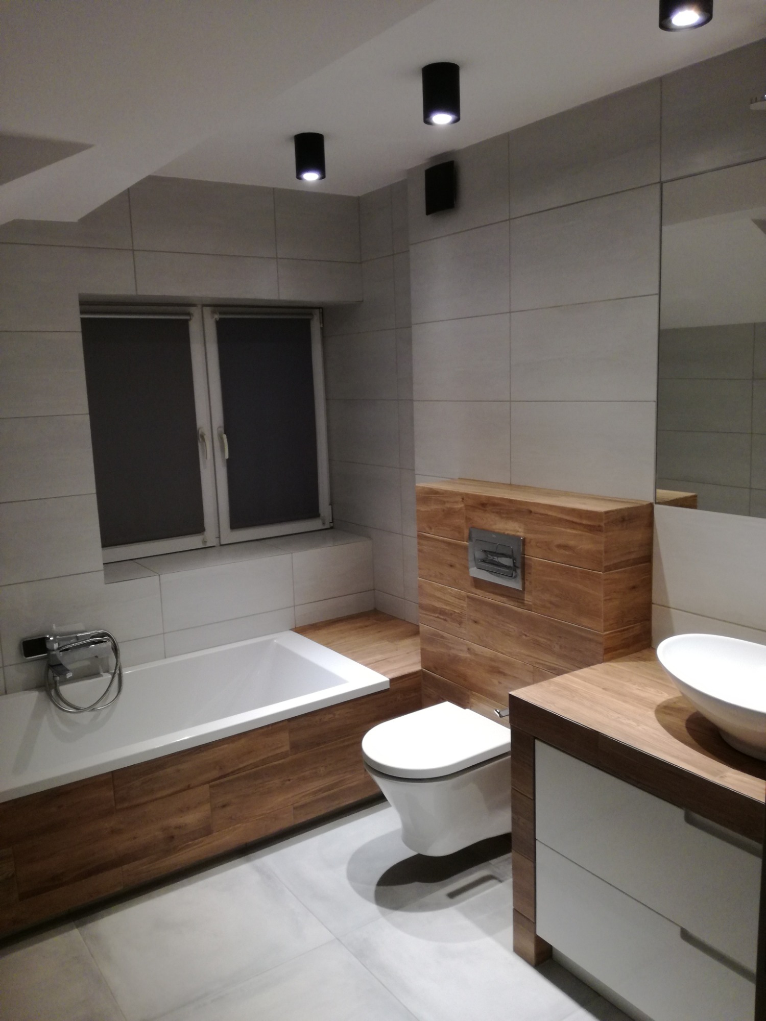 Nowoczesna łazienka z wanną obudowaną drewnopodobnymi panelami, wiszącą toaletą, umywalką nablatową i szarą płytką ścienną.