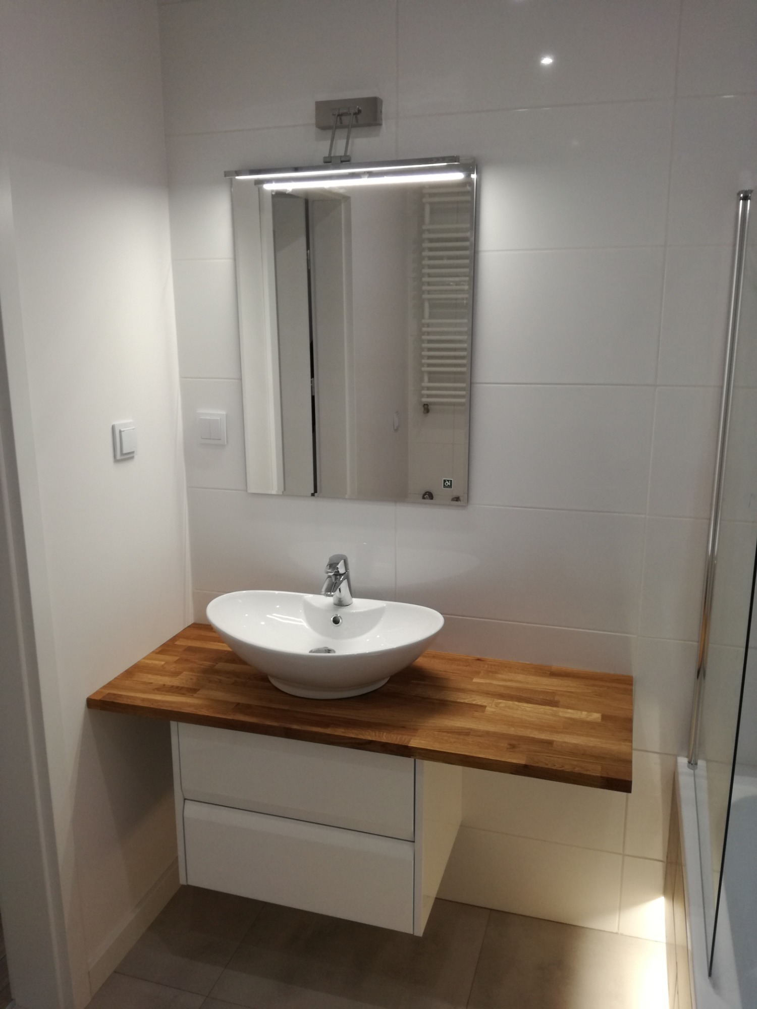 Nowoczesna łazienka z białą umywalką nablatową na drewnianym blacie, podświetlane lustro z grzejnikiem drabinkowym odbijającym się w tafli.