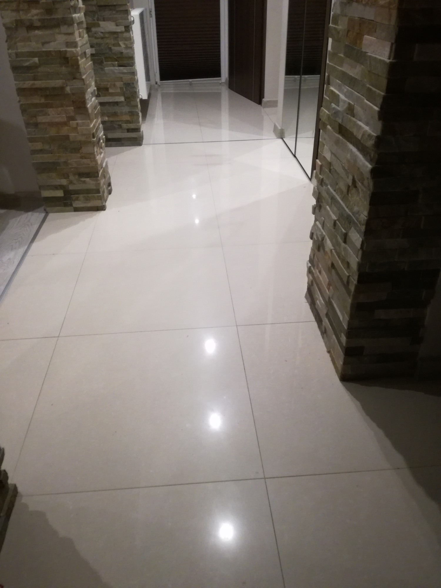 Błyszcząca, beżowa podłoga z płytek w korytarzu, z kolumnami obłożonymi kamieniem dekoracyjnym po bokach, odbicie w lustrzanej szafie w tle.