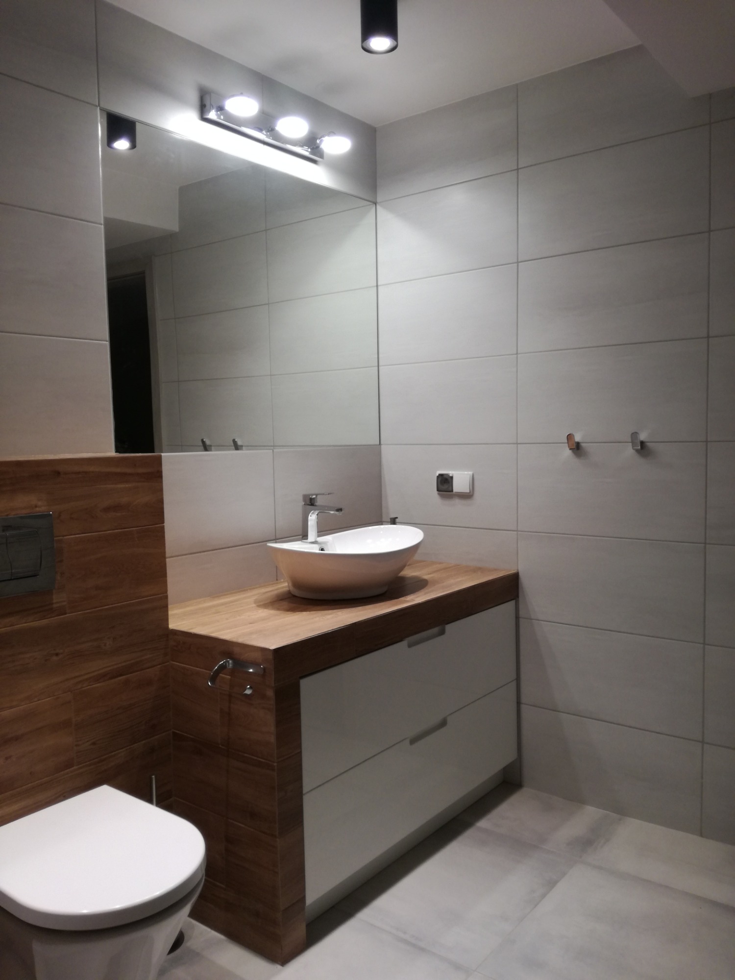 Nowoczesna łazienka z drewnianymi akcentami: blat umywalkowy z drewna, biała umywalka nablatowa, szare płytki ścienne i podłogowe, oraz wisząca toaleta.