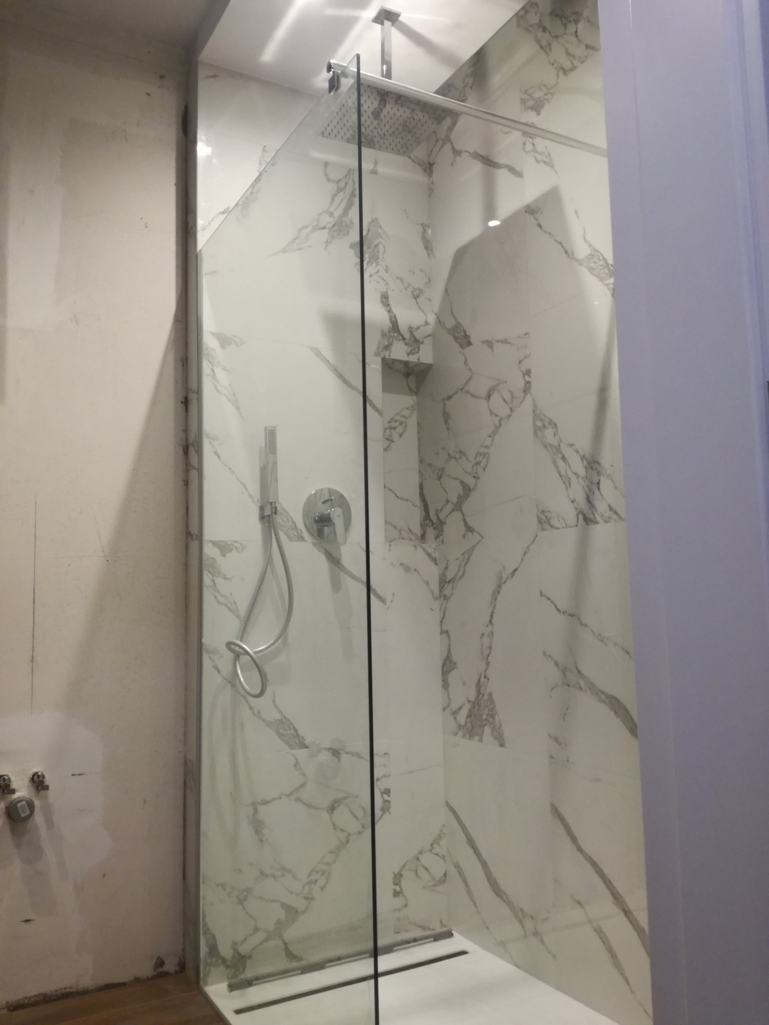 Nowoczesna kabina prysznicowa z deszczownicą i baterią podtynkową na ścianie wyłożonej białymi płytkami imitującymi marmur, widok na ścianę w trakcie prac wykończeniowych.