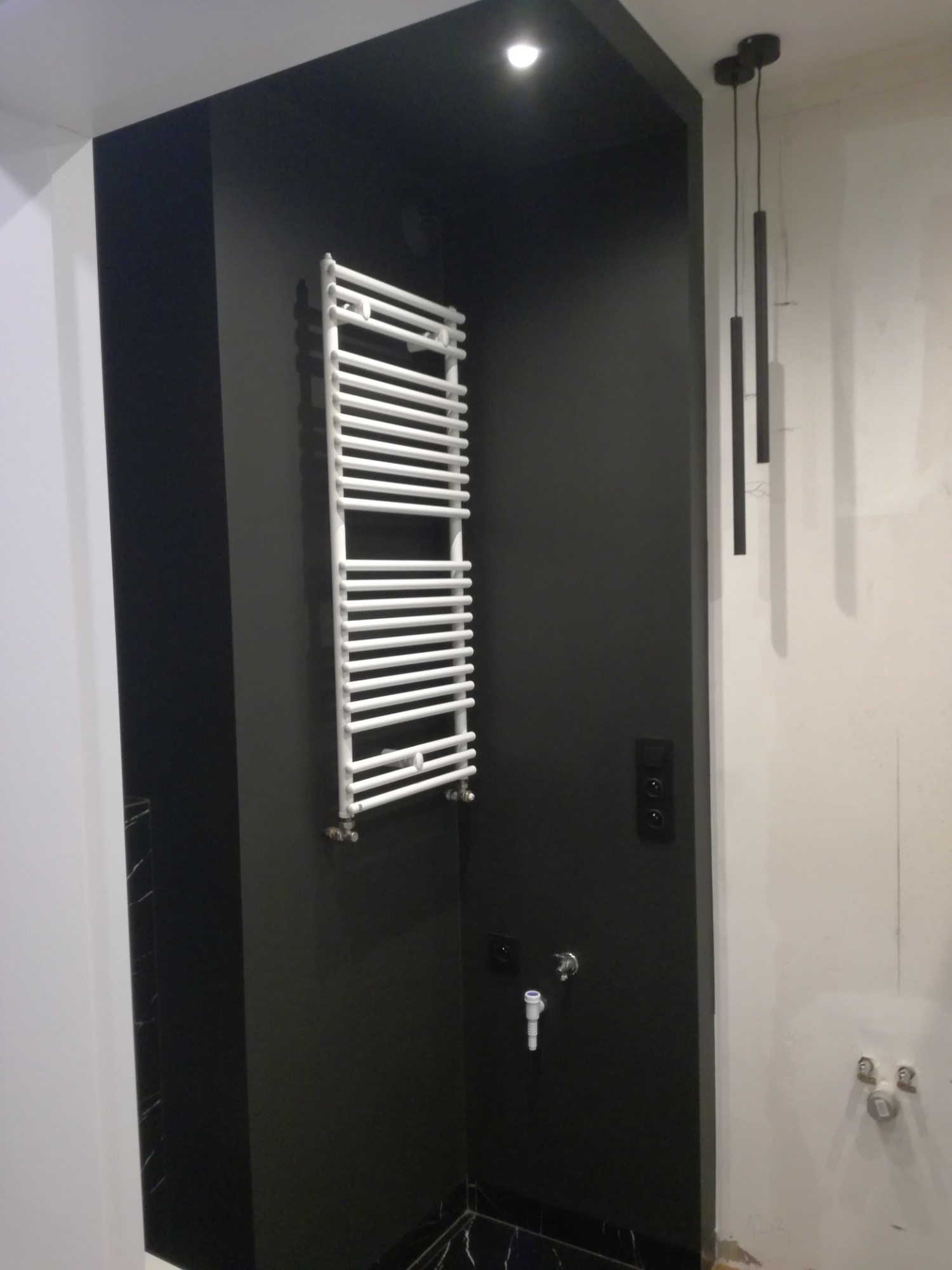 Biały grzejnik drabinkowy na czarnej ścianie w łazience, z widocznymi przyłączami i czarnymi włącznikami światła, obok surowa biała ściana z wystającymi rurami i wiszącymi czarnymi lampami.