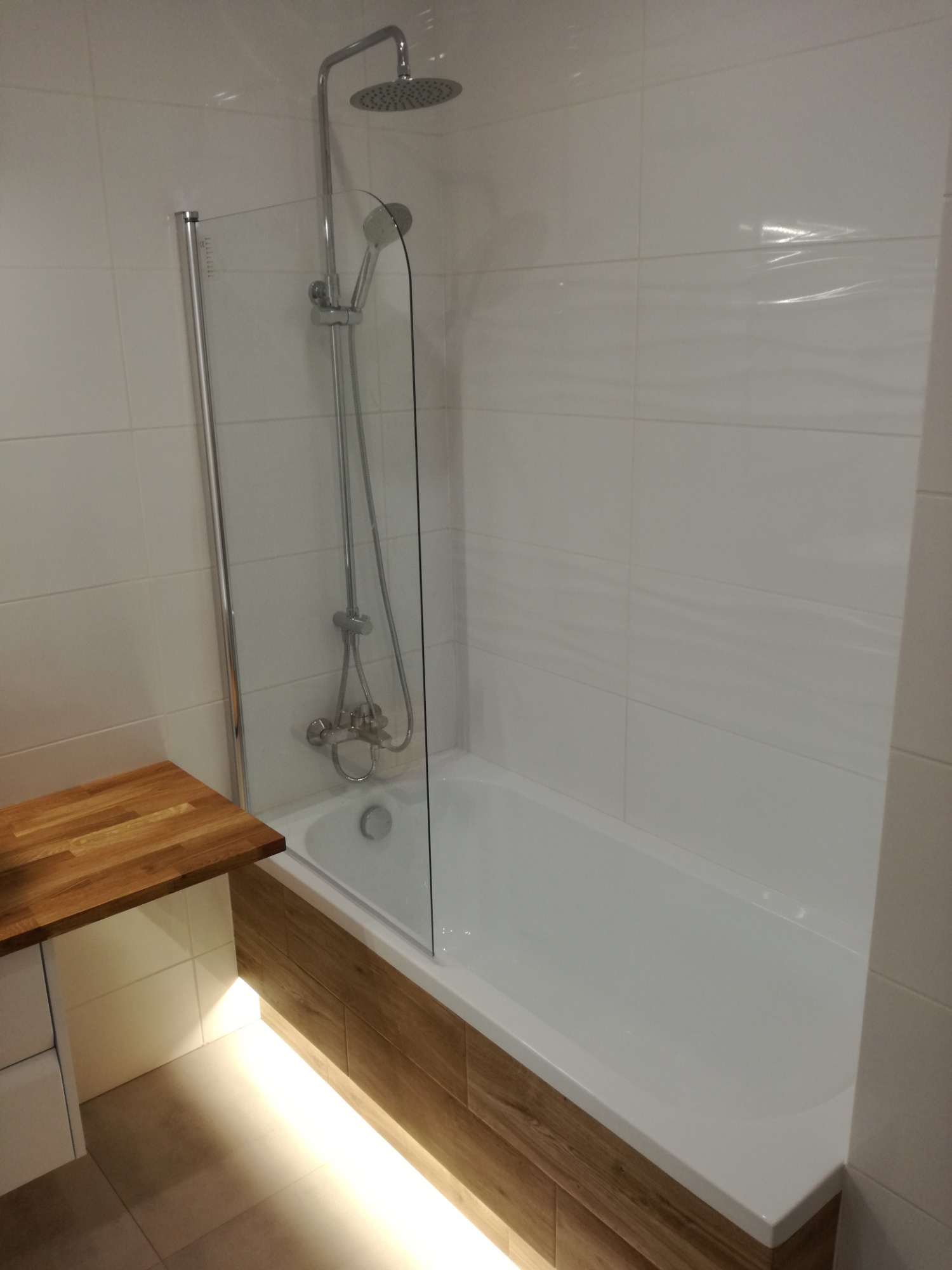 Nowoczesna, wykończona łazienka z białą wanną obudowaną panelami imitującymi drewno, szklanym parawanem nawannowym i chromowaną armaturą prysznicową; dodatkowe oświetlenie LED pod wanną.